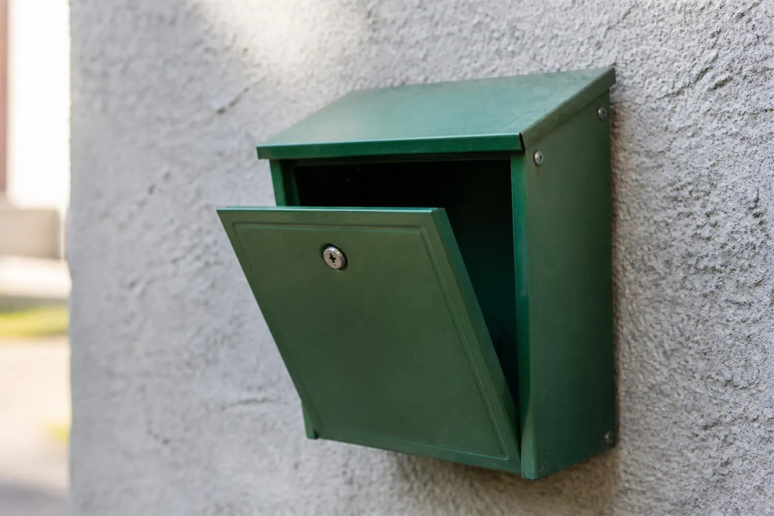 Une boîte aux lettres métallique verte entrouverte et vide, accrochée à un mur de crépi gris.